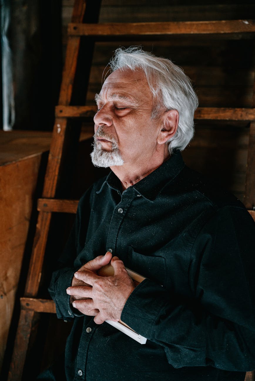 An elderly man with weathered hands carefully carves intricate designs into wooden wind chimes hanging from a tree.